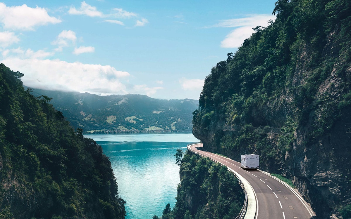 Camping-car profilé Bavaria roulant sur une route de montagne au bord d'un lac.
