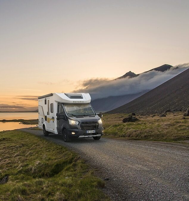 Camping-car profilé étroit Bavaria sur un chemin au bord d'un lac, en fin de journée.