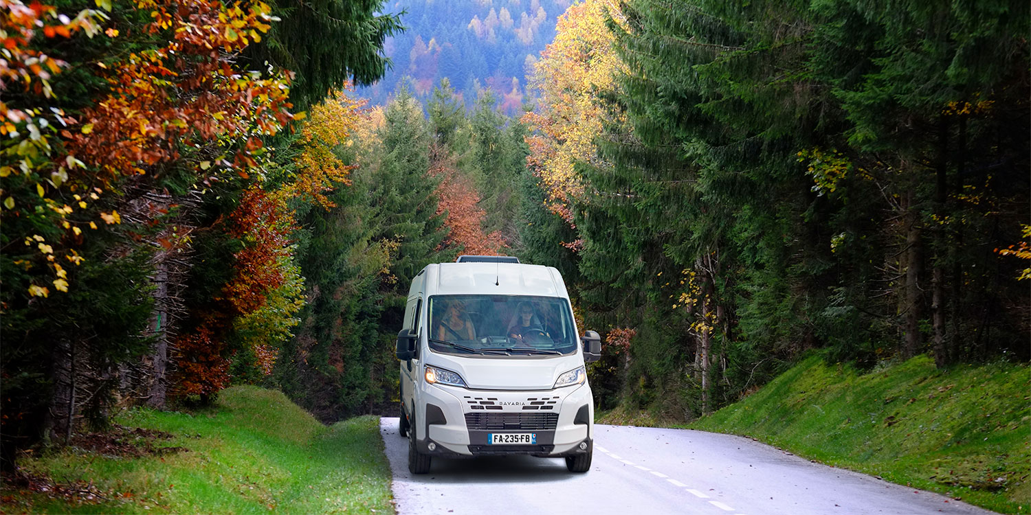 Vue de face d'un fourgon aménagé Bavaria sur une route de montagne en lisière de forêt
