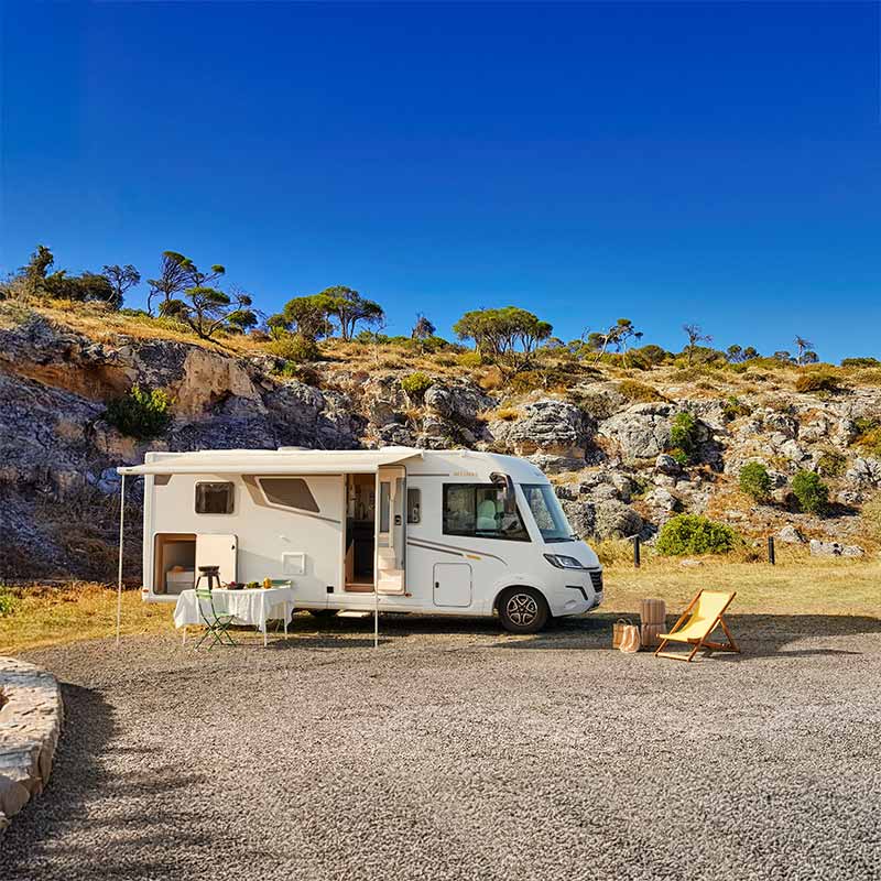 Camping-car intégral Bavaria Intense, installé à l'étape avec store banne, table et chaises dépliés, entouré de roches et de verdures typiquement méditerranéennes.