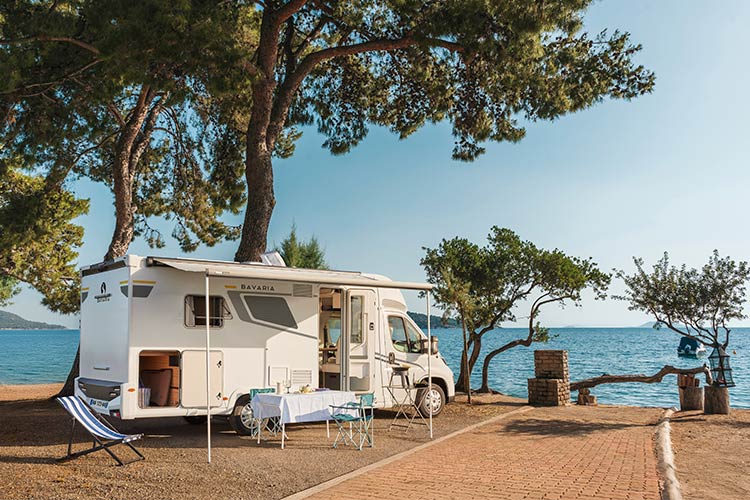 Vue d'un camping-car profilé Bavaria de côté, installé à l'étape dans une aire de camping des Landes, entre les pins et vue mer.