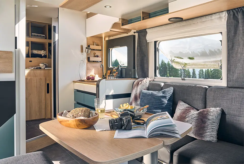 Intérieur d'un camping car profilé étroit avec banquette en tissu gris, table en bois et rangements haut